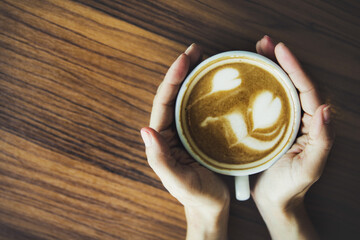 Top view women's hands holding one cup of latte coffee on a brown wood floor : Coffee with beautiful flower motifs by a professional barista.
