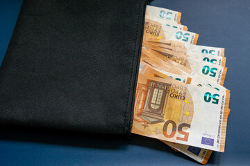 Schwarze Geldtasche mit einem Hundert Euro Schein und mehreren Fünfzig Euro Banknote, vor blauen Hintergrund