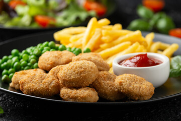 Fried crispy chicken nuggets with ketchup, french fries and green peas in black plate