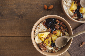 Oatmeal with pineapple, banana and chocolate granola in beige plates on a brown wooden table. A hearty healthy breakfast. A place for text.