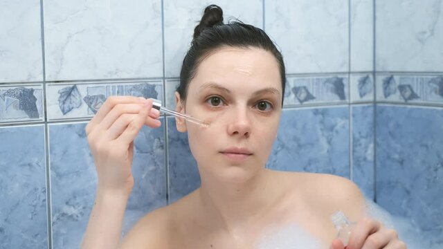 Young woman is applying hyaluronic acid on face skin taking a bath with foam. Rejuvenating and moisturizing, regenerating treatment at home. Wellness, body care and self love concept.