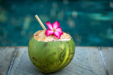 Obraz na płótnie Canvas Green young coconut close up with bamboo straw and tropical pink flower on the edge of swimming pool