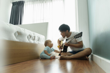 Asian Father and Cute little boy son sitting on wood floor singing and playing acoustic guitar...