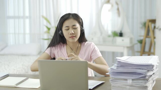 A woman, an office worker or an Asian student wearing black headphones, sitting at work or studying online, is stressed and worried. In front of the laptop It has a lot of documents. Placed on the tab