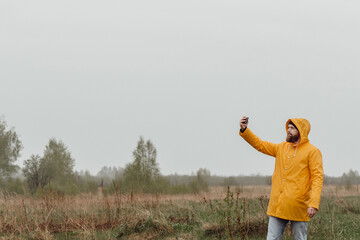 A bearded man in a raincoat on the background of nature.  Man takes pictures by smartphone. Mobile photography.