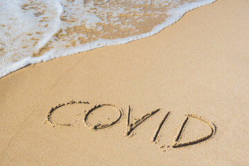 Covid written on the sand of a beach with wave washing the word, erasing or canceling it during Coronavirus summer, Covid free, safe beach, vaccine. Covid-19 immunity passport or pass, bathing season