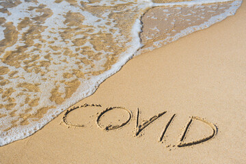 Covid written on the sand of a beach with wave washing the word, erasing or canceling it during Coronavirus summer, Covid free, safe beach, vaccine. Covid-19 immunity passport or pass, bathing season