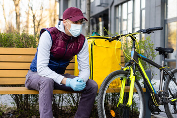 Obraz premium Young courier in medical mask delivering food with yellow thermal backpack, riding a bicycle in the city. Food delivery service concept