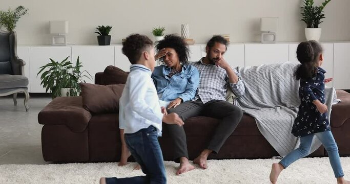 Tired helpless African ethnicity couple sit on sofa feel exhaustion while noisy hyperactive little children running around. Upbringing difficulties, misbehaved son and daughter, ADHD problem concept