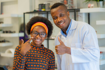 Happy cheerful African teenage girl looking for stylish glasses with qualified African-American optician in optical shop.