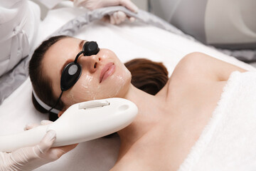 Smiling woman enjoying ELOS skin rejuvenation facial. Female with cooling gel on face and laser protection glasses, lying therapy table at beauty clinic, treating skin from blemishes, doing facelift