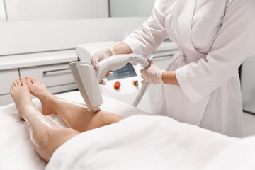 Close up of laser leg hair removal, beautician at spa beauty clinic using hardware cosmetology device for depilation procedure, client legs covered with numbing gel to reduce itching from laser waves