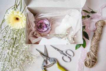 Pink decorative candle in a white gift box on a white table. Nearby are the florist's tools and flowers