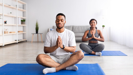 Black guy meditating with closed eyes, sitting in lotus pose, doing namaste gesture, practicing yoga with his girlfriend - obrazy, fototapety, plakaty