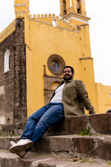 Fototapeta na wymiar Young mexican bearded guy with a jacket sitting on stone steps in a franciscan convent in Mexico. Full body portrait.