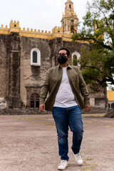 Fototapeta na wymiar Young bearded mexican man wearing green jacket and face mask walking in front of a church in Mexico. Full Body Portrait