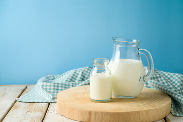 Milk jug and milk bottle on wooden table with tablecloth and log tray over blue background. Kitchen mock up for design
