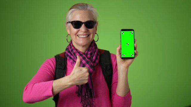 Portrait of happy smiling mature woman traveler with backpack pointing to smartphone with green screen