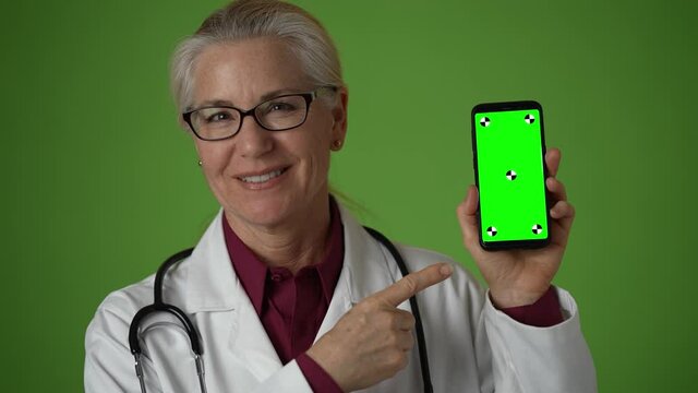 Closeup portrait of happy smiling female doctor holding smartphone on green screen and pointing to it.