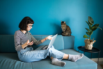 Young woman working with laptop and with a cat sitting in modern living room at home. Woman with her cat spending time together. Education online. Working from home in quarantine lockdown.