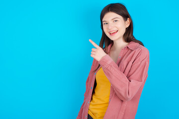 young beautiful Caucasian woman wearing pink jacket over blue wall pointing away and smiling to you. Look over there!