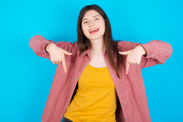 Fototapeta premium young beautiful Caucasian woman wearing pink jacket over blue wall with positive expression, points down with both index fingers, has broad interested smile. Look there, please.