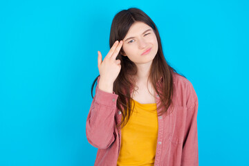 Unhappy young beautiful Caucasian woman wearing pink jacket over blue wall imitates gun shoot makes suicide gesture keeps two fingers on temples.