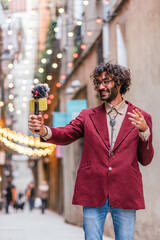 attractive caucasian man recording himself in the streets of Barcelona. He is holding a gimbal with a yellow mobile phone and a microphone. He is dressing casual with blue jeans and red jacket.