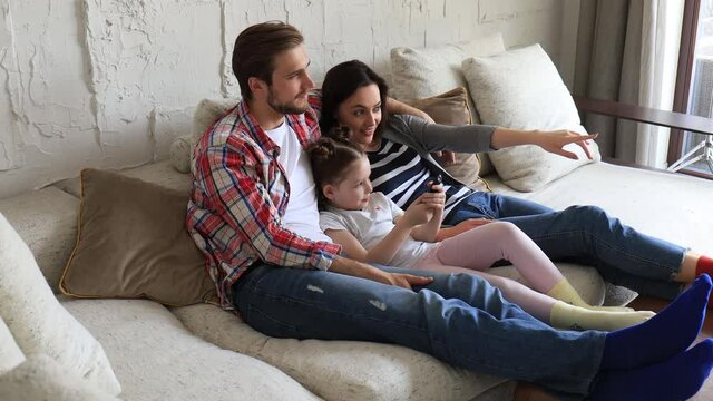 Beautiful happy young parents and their little daughter watching TV and smiling while sitting on sofa at home.