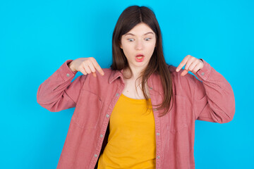 Amazed young beautiful Caucasian woman wearing pink T-shirt over blue wall points down with fore...