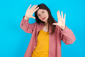 Portrait of smiling young beautiful Caucasian woman wearing pink T-shirt over blue wall looking at camera and gesturing finger frame. Creativity and photography concept.