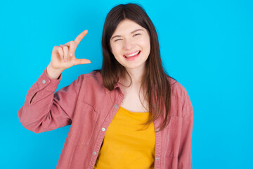 Fototapeta premium young beautiful Caucasian woman wearing pink jacket over blue wall smiling and gesturing with hand small size, measure symbol.