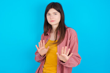 Afraid young beautiful Caucasian woman wearing pink jacket over blue wall, makes terrified expression and stop gesture with both hands saying: Stay there. Panic concept.