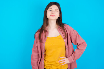 Studio shot of cheerful young beautiful Caucasian woman wearing pink jacket over blue wall keeps hand on hip, smiles broadly.