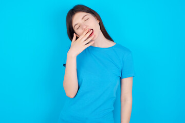 Sleepy young beautiful Caucasian woman wearing blue T-shirt over blue wall yawning with messy hair, feeling tired after sleepless night, yawning, covering mouth with palm.