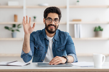 Happy arab man waving hand and smiling to camera, sitting at workplace at home. Modern remote...