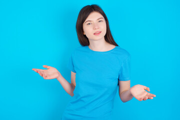 young beautiful Caucasian woman wearing blue T-shirt over blue wall looks uncertain shrugs...