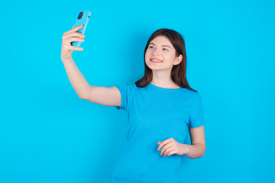 Portrait of a young beautiful Caucasian woman wearing blue T-shirt over blue wall taking a selfie to send it to friends and followers or post it on his social media.