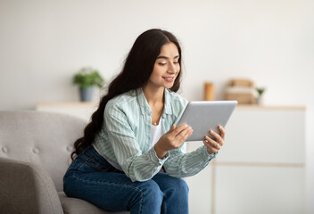 Cheerful Indian lady sitting in armchair with tablet computer, studying remotely or having online...