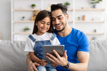 Smiling dad and daughter sitting on couch, using digital tablet