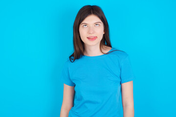 Obraz premium Funny young beautiful Caucasian woman wearing blue T-shirt over blue wall makes grimace and crosses eyes plays fool has fun alone sticks out tongue.