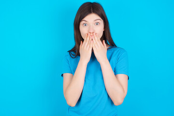 young beautiful Caucasian woman wearing blue T-shirt over blue wall keeps hands on mouth, looks with eyes full of disbelief, being puzzled with amount of work