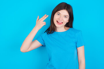 Fototapeta premium young beautiful Caucasian woman wearing blue T-shirt over blue wall foolishes around shoots in temple with fingers makes suicide gesture. Funny model makes finger gun pistol