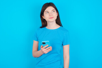 young beautiful Caucasian woman wearing blue T-shirt over blue wall holds mobile phone uses high speed internet and social networks has online communication. Modern technologies concept