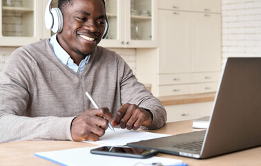 Adult happy smiling African American black male student wearing headphone sit at kitchen table,...