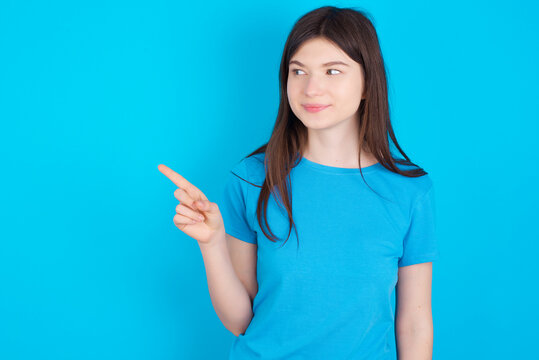 young beautiful Caucasian woman wearing blue T-shirt over blue wall points at copy space and advertises something, advices best price.