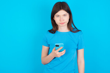 Upset dissatisfied young beautiful Caucasian woman wearing blue T-shirt over blue wall uses mobile software application and surrfs information in internet, holds modern mobile hand