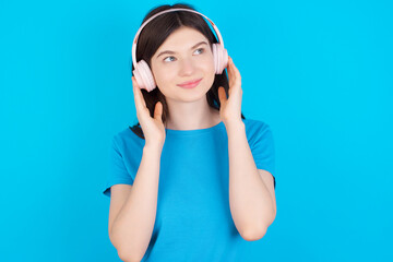 Naklejka premium young beautiful Caucasian woman wearing blue T-shirt over blue wall wears stereo headphones listens music concentrated aside. People hobby lifestyle concept