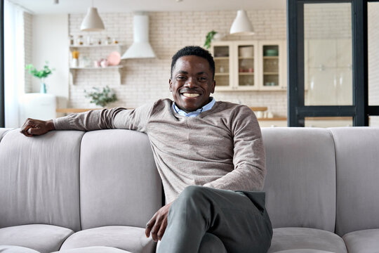 Young adult happy cheerful smiling African American business man with toothy smile relaxing posing sitting on comfortable sofa at home modern living room looking at camera. Work life balance concept.