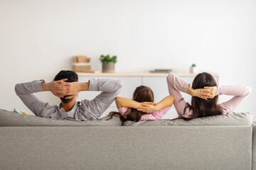 Leisure time. Back view of relaxed parents and child with arms behind head sitting on couch and...